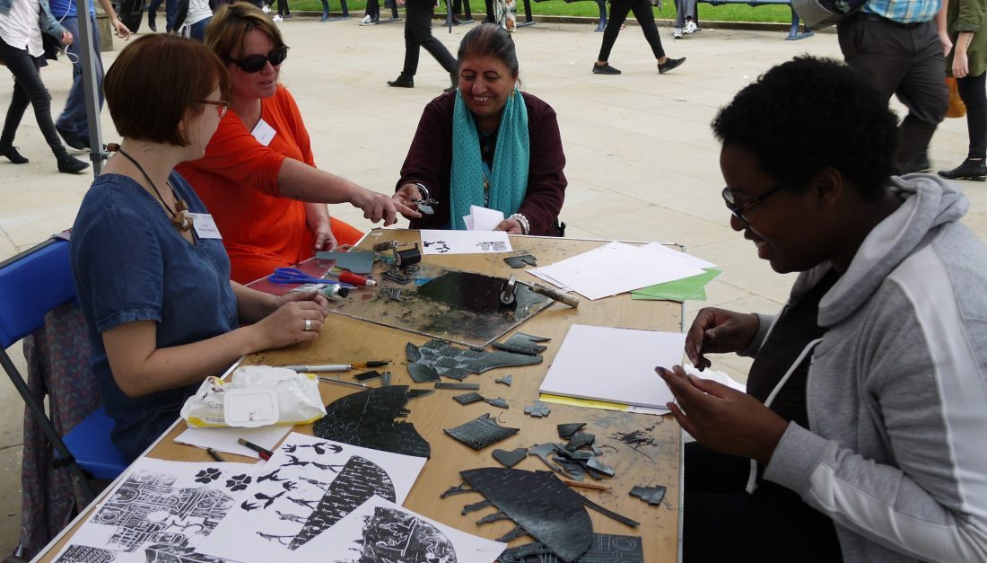 Three women interact and talk to the artist around a table whilst linoprinting.